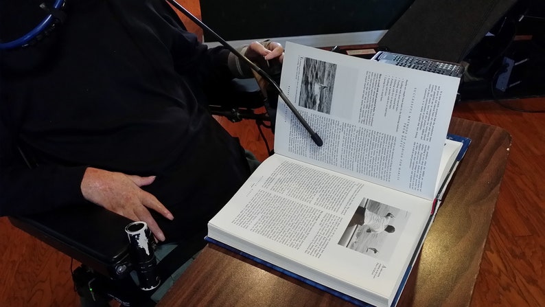 May include: A person in a wheelchair is reading a large, open book with a black light attached to a flexible arm. The book has a black and white photo of a bird on the water on the left page and a black and white photo of a person on the right page.