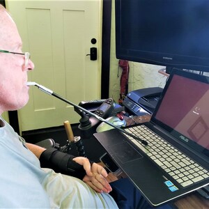 May include: A person using a mouth stick to control a laptop computer. The laptop is on a desk with a black surface. The laptop has a black case and a white keyboard. The screen is displaying a loading screen with the word "ASUS" on it.
