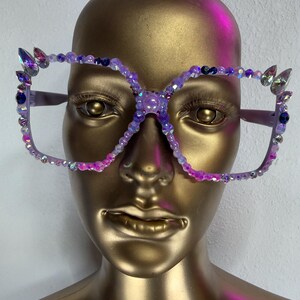 May include: Lavender-colored, oversized eyeglasses with a square frame. The frames are adorned with iridescent beads and crystals in shades of purple, pink, and blue. The bridge of the glasses features a decorative cluster of beads and crystals.