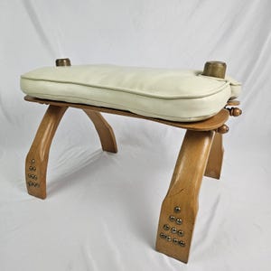 May include: A footstool featuring a cream-colored padded cushion top and a light brown wooden frame. The four curved wooden legs are accented with small, decorative metal studs at the base, and two brass-colored cylindrical caps are visible on top.