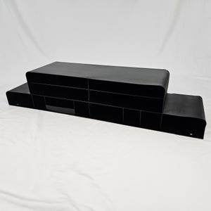 May include: A matte black metal desk organizer featuring a tiered design with multiple rectangular compartments. The structure consists of a long, low base section with several dividers and a raised, wider upper shelf with two main storage sections.