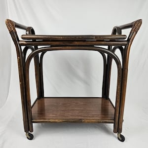 May include: A dark brown rattan serving cart featuring two rectangular tiers with a smooth wooden surface on the bottom shelf. The frame is constructed from curved bamboo-style poles with woven detailing at the top handles and four small wheels.