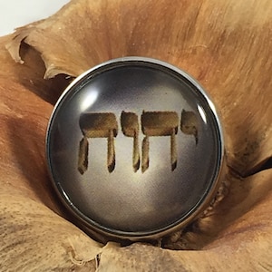 May include: A silver-toned metal button with a clear glass top featuring a gold Hebrew inscription. The inscription reads "Shema Yisrael" which translates to "Hear O Israel".