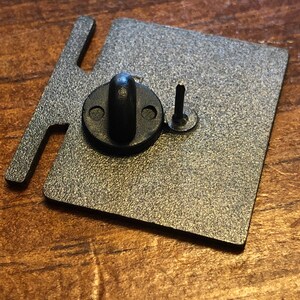 Op de afbeelding: Een getextureerde, metaalkleurige metalen pinrug met een zwarte rubberen sluiting en een pin. De pinrug heeft een rechthoekige vorm met een lip aan de linkerkant. De rug is ontworpen om een pin vast te zetten.