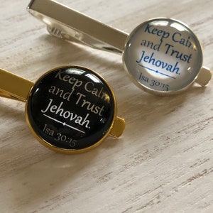 May include: Two tie clips with round glass inserts. The tie clips are gold and silver. The glass inserts have black and white text that reads "Keep Calm and Trust Jehovah. Isa 30:15".