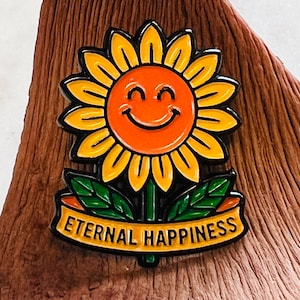 Puede incluir: Un pin de esmalte de girasol alegre con una cara sonriente, pétalos amarillos y un centro naranja. El pin presenta hojas verdes y una pancarta que dice "ETERNAL HAPPINESS" en letras negras. Sobre un fondo de madera.