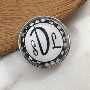 May include: A white round button with a black and white checkered border. The button has a black monogram of the letters "DL" in a script font.