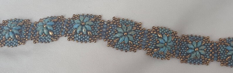 May include: A beaded bracelet with a repeating pattern of blue and gold flowers. The bracelet is made with small seed beads and features a delicate, intricate design.