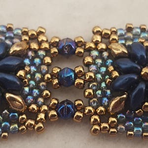 May include: A close-up of a beaded bracelet with a square design featuring two blue flowers with gold accents. The beads are iridescent blue, gold, and dark blue.