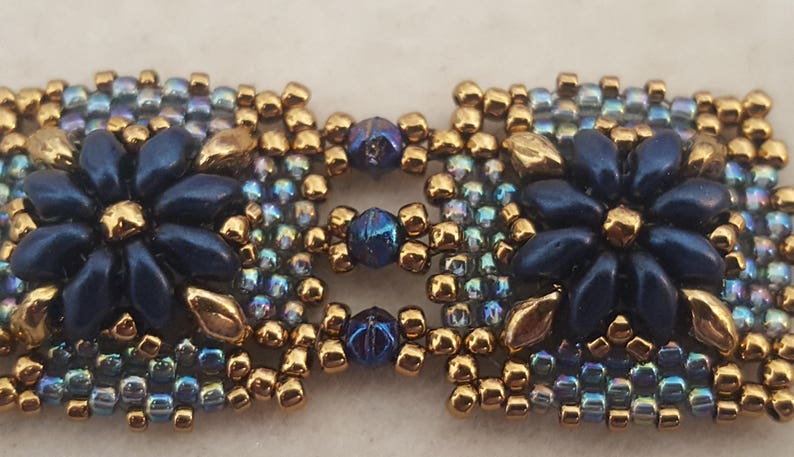 May include: A close-up of a beaded bracelet with a square design featuring two blue flowers with gold accents. The beads are iridescent blue, gold, and dark blue.