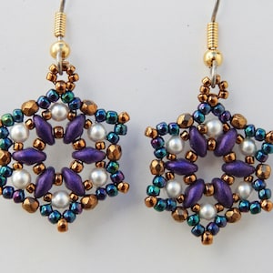 Può includere: Un paio di orecchini con un design a fiocco di neve. I fiocchi di neve sono fatti di perline viola, dorate e verdi. Gli orecchini sono appesi a ganci d'argento.