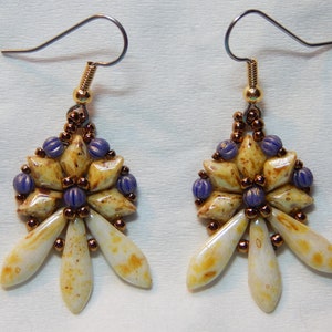 May include: A pair of dangle earrings with a white and brown flower design. The flower has a center of purple beads and is surrounded by smaller brown beads. The earrings are attached to silver hooks.