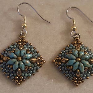 May include: A pair of earrings with a square-shaped design featuring a turquoise blue flower with gold accents. The earrings are made with small beads and have silver hooks.