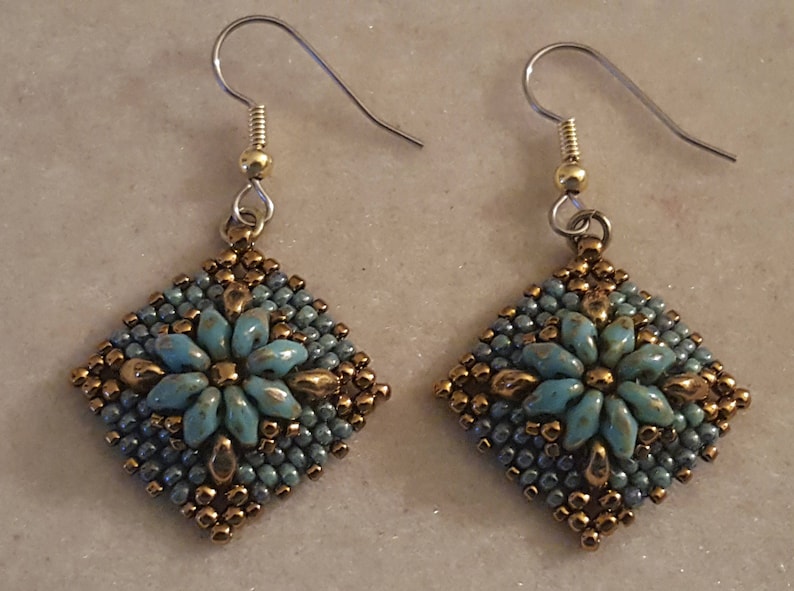 May include: A pair of earrings with a square-shaped design featuring a turquoise blue flower with gold accents. The earrings are made with small beads and have silver hooks.