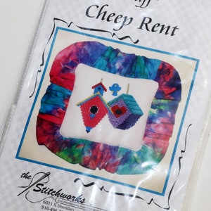 Puede incluir: Una almohada blanca con un borde de tela de teñido anudado de colores. La almohada presenta un diseño de punto de cruz de dos casas para pájaros con acentos rojos y morados. El empaque incluye el texto "Fibers-n-Stuff Cheep Rent" y "the Stitchworks".