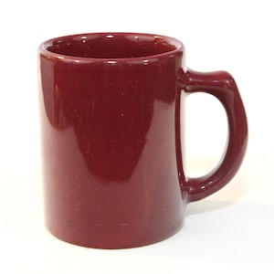 May include: A burgundy ceramic mug with a rounded handle. The mug has a speckled texture and a glossy finish. The handle is a solid, curved shape, and the mug is cylindrical. The mug is empty and sits on a white background.
