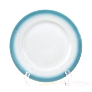 Puede incluir: Un plato blanco con un borde azul turquesa. El plato es redondo y tiene un borde ligeramente elevado. El color azul turquesa se desvanece en el centro blanco del plato. El plato se exhibe en un soporte de plástico transparente.