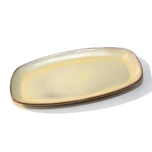 May include: A rectangular ceramic serving platter with rounded corners. The platter has a light yellow center with a gradient to a darker, olive-green edge. A thin, brown line borders the edge of the platter. The background is white.