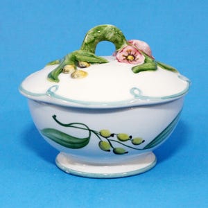 May include: White ceramic sugar bowl with a lid, featuring hand-painted floral and leaf designs. The lid has a green handle and pink flowers. The bowl has a light blue trim and a matching base. The design includes green leaves and yellow berries.