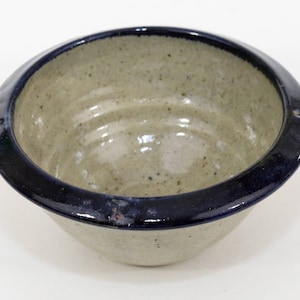 May include: A small, white ceramic bowl with a dark blue rim. The bowl has a speckled glaze and a slightly textured surface.