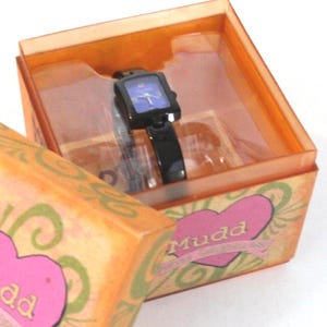 Könnte beinhalten: Eine schwarze Mudd-Uhr mit quadratischem blauem Zifferblatt wird in einer orangefarbenen Schachtel präsentiert. Die Uhr hat ein schwarzes Armband und ein quadratisches Zifferblatt. Die Schachtel ist mit rosa Herzen und dem Wort "Mudd" verziert.