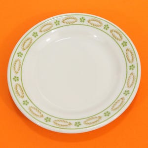 May include: A white ceramic plate with a decorative border. The border features a repeating pattern of wheat-like shapes and small green flowers. The plate has a simple, classic design.