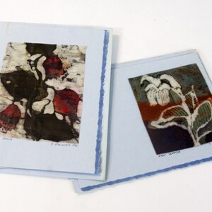 May include: Two floral art prints on light blue paper. One print features dark red and black flowers, the other a white and green flower. The prints are titled "Tajo Della" and "Lady Slipper".