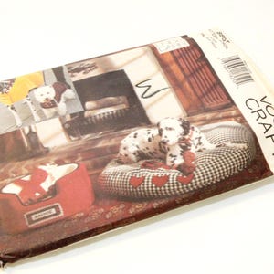 May include: Vogue Craft sewing pattern for dog beds. The pattern features a Dalmatian dog on a checkered bed with red heart accents. The pattern also shows a red dog bed with the name "Annie" on it. The pattern's cover has a vintage aesthetic.