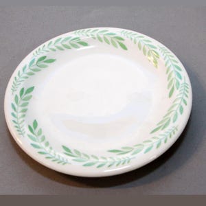 Puede incluir: Plato de cerámica blanco con un patrón de hojas verdes alrededor del borde. Las hojas están pintadas al estilo acuarela, creando una estética delicada y natural. Este plato es adecuado para servir alimentos.