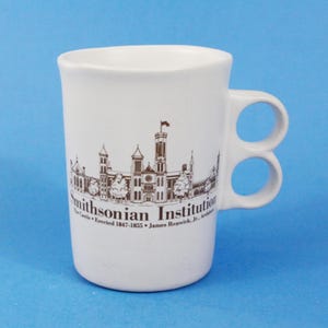 Può includere: Tazza in ceramica bianca con un'illustrazione marrone dell'edificio dello Smithsonian Institution e il testo "Smithsonian Institution The Castle - Erected 1847-1855 - James Renwick, Jr., Architect". La tazza ha due manici.
