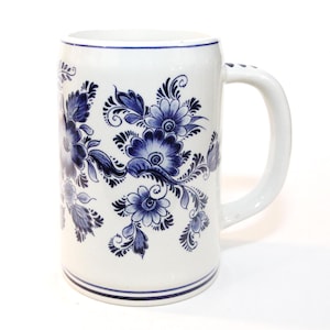 May include: White ceramic beer stein with a blue floral design. The mug features a large handle and a cylindrical shape. The blue floral pattern is hand-painted and covers most of the mug's surface. A blue stripe accents the top and bottom.