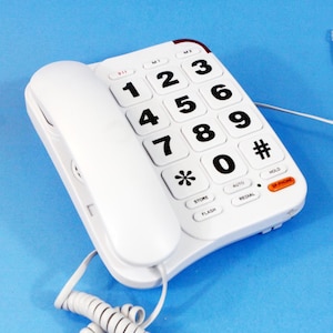 Könnte beinhalten: Weißes schnurgebundenes Telefon mit einem großen Tastenfeld und einem Hörer. Das Telefon hat Tasten zum Speichern, Blitz, Auto, Halten und Wählen.