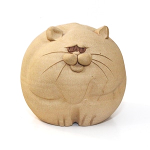 May include: A round, beige ceramic cat figurine with a smiling face and whiskers. The cat has small, upright ears and a slightly flattened appearance. The eyes and nose are a darker brown color.