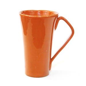 May include: An orange ceramic mug with a distinctive handle. The mug has a glossy finish and a tapered shape. The handle is angular and attached to the side. The mug is empty and set against a white backdrop. The mug's height is approximately 15 cm.