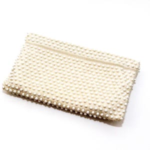 May include: A rectangular off-white clutch bag featuring a dense, uniform pattern of small, raised white beads covering the entire exterior surface. The bag has a structured, flat design with a horizontal seam detail across the upper portion.