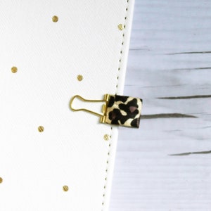 May include: A gold binder clip with a leopard print ribbon attached. The binder clip is on a white background with gold polka dots.