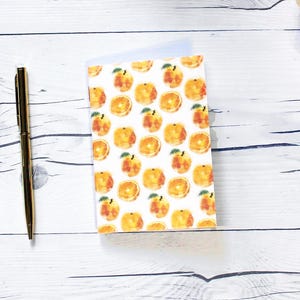 Peut inclure: Un petit carnet avec un fond blanc et un motif répété de tranches d'orange. Les tranches d'orange sont peintes dans un style aquarelle avec des feuilles vertes.