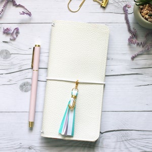 May include: White leather notebook with a white ribbon and a turquoise tassel with a gold letter J charm.