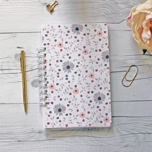 May include: A white spiral bound notebook with a floral pattern in shades of pink, gray, and blue. The notebook is on a white wooden surface.