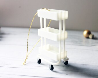 May include: A miniature white rolling cart with three shelves and black wheels. A gold string is attached to the top. The cart is a decorative item, possibly an ornament, and is set against a blurred background with gold ornaments.