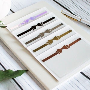 May include: Five clear plastic cards with a white background and a metallic bow design in different colours. The bows are in shades of purple, black, grey, gold, and brown.