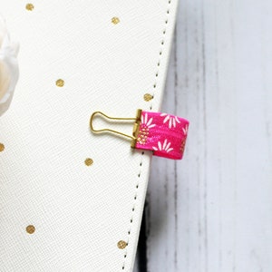 May include: A gold binder clip with a pink elastic band with a white daisy pattern. The elastic band is attached to the binder clip.