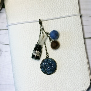 May include: A gold chain with a small glass bottle, a feather, and three felt balls in blue and brown. The bottle has a label that says "Ink". The chain is attached to a round, blue, and gold charm.