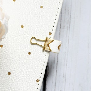 Peut inclure: Un trombone doré avec un ruban à motif chevron blanc et or attaché. Le trombone est sur une surface blanche avec des pois dorés.