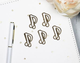 May include: Five gold metal paper clips shaped like musical notes on a white surface with gold glitter.