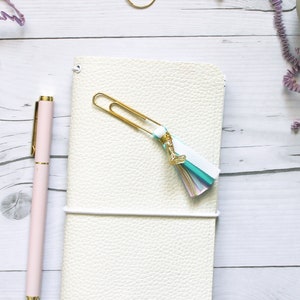 May include: A white leather notebook with a gold paper clip and a colourful tassel. The notebook is closed and has a white elastic band.