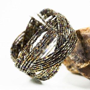 May include: A woven beaded bracelet with a multi-colored iridescent finish. The bracelet is made of small, round beads in shades of brown, gold, and silver. The bracelet is lying on a rough, brown rock.