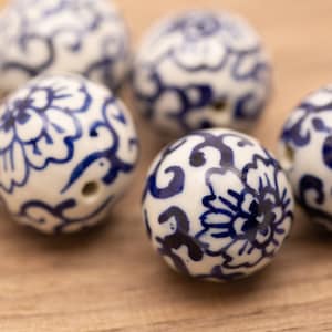 May include: A collection of six white ceramic beads with blue floral patterns. The beads are round and have a hole in the center for stringing.