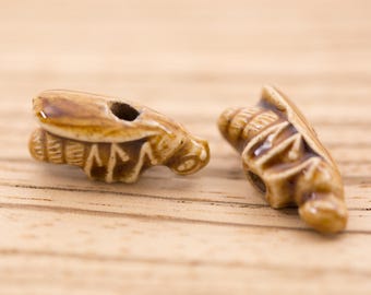 May include: Two ceramic beads shaped like bugs, one brown and one light brown, with intricate details. The beads are on a wooden surface.