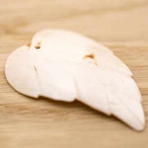 May include: A carved, off-white feather pendant with a small hole at the top. The feather has detailed lines and a slightly irregular shape, with brown spots. The pendant rests on a light brown wooden surface.